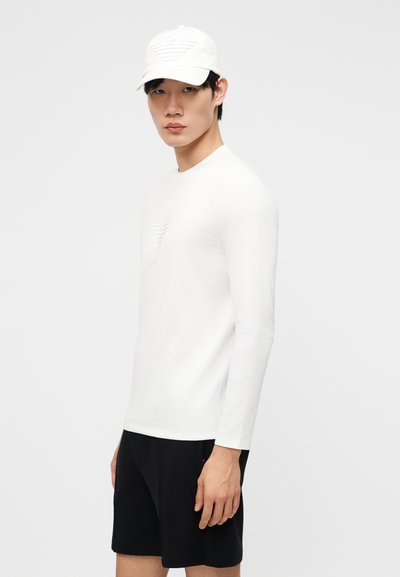 Νεαρός άνδρας που φοράει λευκό πουκάμισο με μακριά μανίκια και καπέλο, με μαύρο σορτς, στέκεται μπροστά σε απλό ανοιχτό γκρι φόντο.