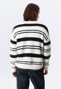 Personne avec des cheveux bruns mi-longs portant un pull blanc duveteux avec des rayures horizontales noires, de dos contre un fond uni.