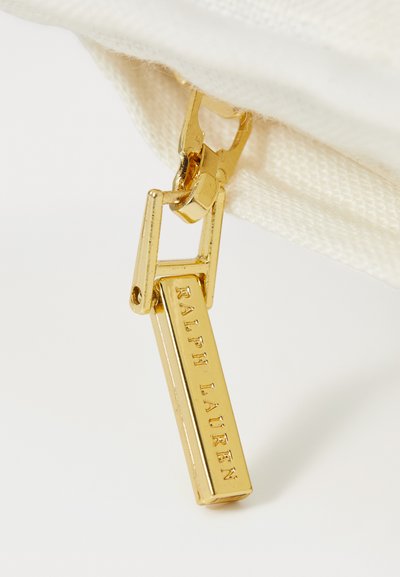 Gouden rits trektong met een rechthoekig label gegraveerd met "RALPH LAUREN." Bevestigd aan een crèmekleurige stoffen tas met een gestructureerd oppervlak.