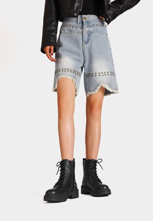 Shorts in denim di un blu chiaro con orli sfrangiati e dettagli in borchie argentate, abbinati a stivali da combattimento neri con lacci e suola chunky.