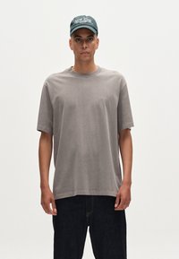 Tricou gri supradimensionat cu mâneci scurte, guler rotund și branding subtil. Asortat cu blugi negri din denim și o șapcă verde de baseball.