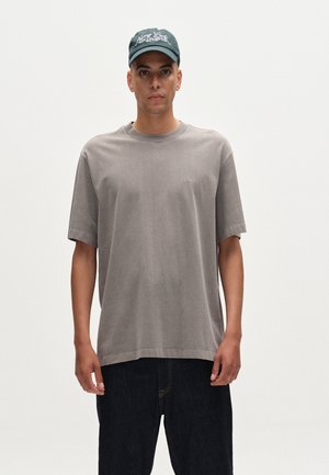 Gray oversized t-shirt with short sleeves, round neck, and subtle branding. Paired with dark blue denim jeans and a green baseball cap.