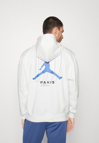 Hvid hættetrøje med logoet Jumpman i blå og teksten "PARIS" på ryggen, med ribbede manchetter og talje.
