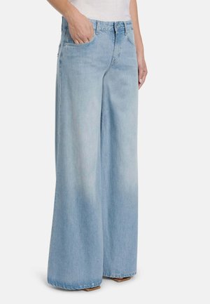 Persona che indossa jeans azzurri a gamba larga con mano in tasca, abbinati a una maglia bianca su uno sfondo semplice.