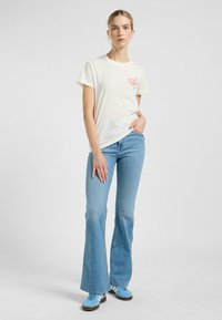 Hvid bomulds-T-shirt med lyserødt hjerteprint, parret med lyseblå flairbukser og blå sneakers, mod en ensfarvet baggrund.
