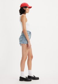 Biely tielko, obnošené modré džínsy, červená šiltovka, čierne mokasíny a biele ponožky; bočný profil ukazuje ležérny letný outfit.