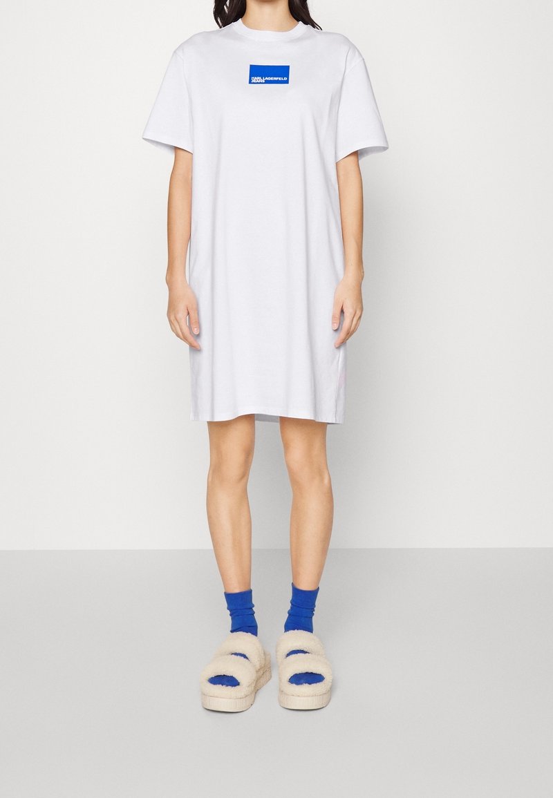 Weißes T-Shirt-Kleid mit einem blauen Logopatch. Das Model trägt beiges, flauschiges Sandalen und blaue Knöchelsocken, mit einem schlichten, lässigen Design.