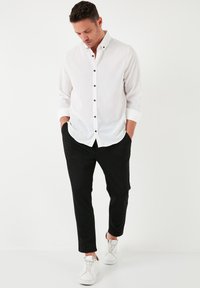 Camisa blanca de botones con un tejido texturizado, botones negros y puños. Combinada con pantalones negros ajustados y zapatillas blancas. Diseño minimalista.