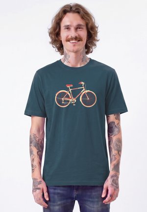 Mann mit lockigem Haar und Tätowierungen lächelt, trägt ein türkisfarbenes T-Shirt mit rotem und beigem Fahrradmotiv und steht vor einem schlichten weißen Hintergrund.