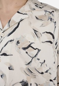 Lett, kremfarget topp i stoff med abstrakt blomsterdesign i beige, blått og svart. Har krage og knappelukking.