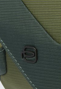 Sac en tissu texturé vert foncé et olive avec un logo noir en accent, présentant une finition lisse et des coutures contrastées.