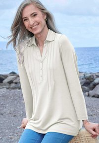 Pullover beige chiaro a maniche lunghe con colletto, dotato di patta con bottoni e pieghe verticali. Indossato sopra jeans blu, con una texture di tessuto morbida.