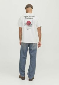 White cotton t-shirt with a graphic print featuring a red circle and mountain lines on the back. Paired with loose blue denim jeans.