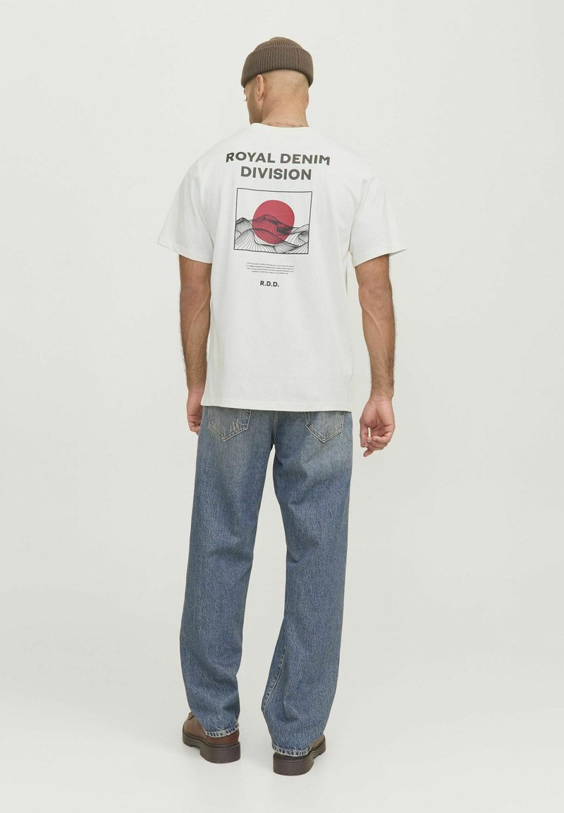 White cotton t-shirt with a graphic print featuring a red circle and mountain lines on the back. Paired with loose blue denim jeans.