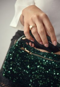 Sac à main vert en sequins avec une fermeture éclair, présentant une surface texturée. Porté par une main portant une bague en or, avec une sangle en cuir lisse.