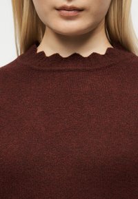 Gros plan d'une personne portant un pull en maille bordeaux avec un col rond festonné, montrant uniquement les lèvres et le menton.