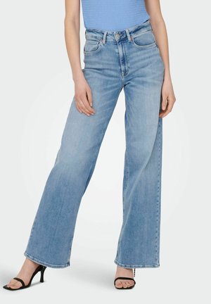 Kvinde iført lyseblå jeans med høj talje og vide ben, en blå ribbet top og sorte åbne hælede sandaler, stående med benene let fra hinanden.