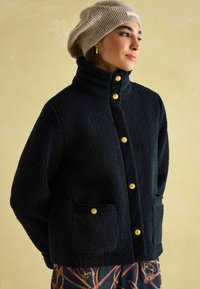 Chlpatá tmavomodrá bunda s vysokým golierom, zlatými gombíkmi a bočnými vreckami. Nosí sa s béžovým pleteným baretom. Hladké textílie kontrastujú s textúrou.
