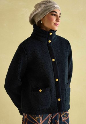 Flauschige marineblaue Jacke mit hohem Kragen, goldenen Knöpfen und Seitentaschen. Getragen mit einer beigen Strickberet. Glatte Stoffakzente setzen einen Kontrast zur Textur.