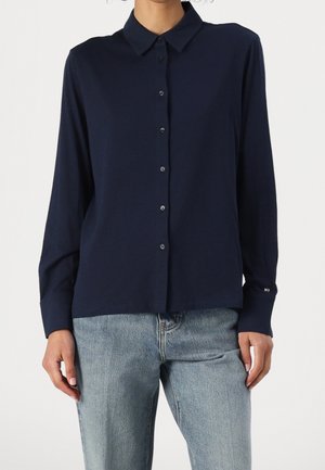 Personne portant une chemise bleu marine à boutons avec manches longues et col, et un jean bleu clair, debout devant un fond uni.