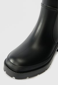 Bottine noire en caoutchouc avec une finition lisse, bout rond et semelle de traction texturée avec des accents nervurés.