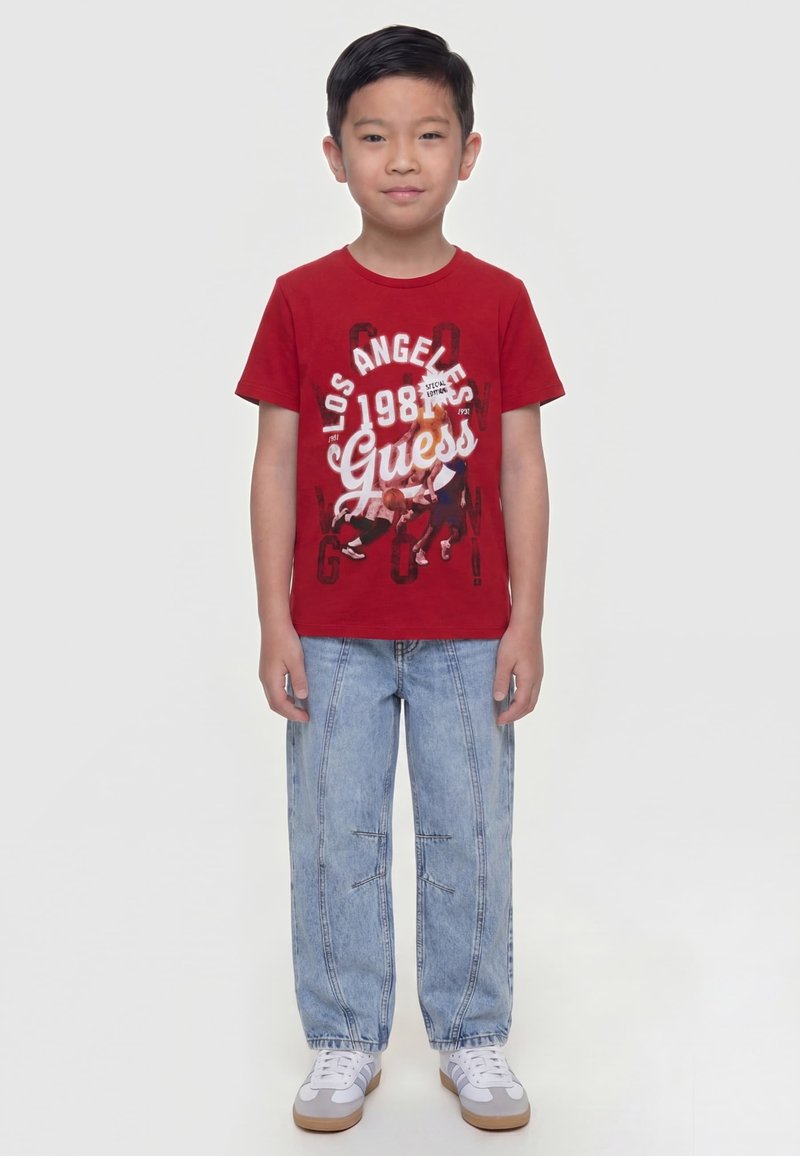 Garçon portant un T-shirt rouge avec un motif graphique et le texte « Los Angeles 1981 Guess », un jean bleu clair et des baskets blanches, debout les mains le long du corps.