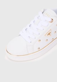 Scarpa da ginnastica bianca con borchie dorate, suola bordata d'oro, lacci bianchi e logo Guess dorato sul lato e sulla linguetta.