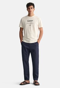 Beige bomulds t-shirt med marineblå logo tekst, parret med mørkeblå bukser og brune sandaler. Outfittet har en afslappet og løs pasform.