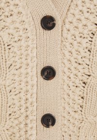 Beige Strickjacke mit strukturiertem Muster, sechs großen dunklen Knöpfen und V-Ausschnitt. Weiches Garn mit sichtbaren Nahtdetails.