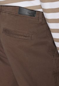 Gros plan sur la poche arrière marron du pantalon et la boucle de ceinture avec l'étiquette noire Jack & Jones, porté avec une chemise beige et blanche à rayures.