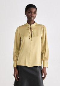 Blusa color oliva chiaro con colletto alla coreana, chiusura frontale con laccetto e maniche lunghe plissettate. Tessuto liscio con una vestibilità comoda, abbinata a una gonna di pelle nera.