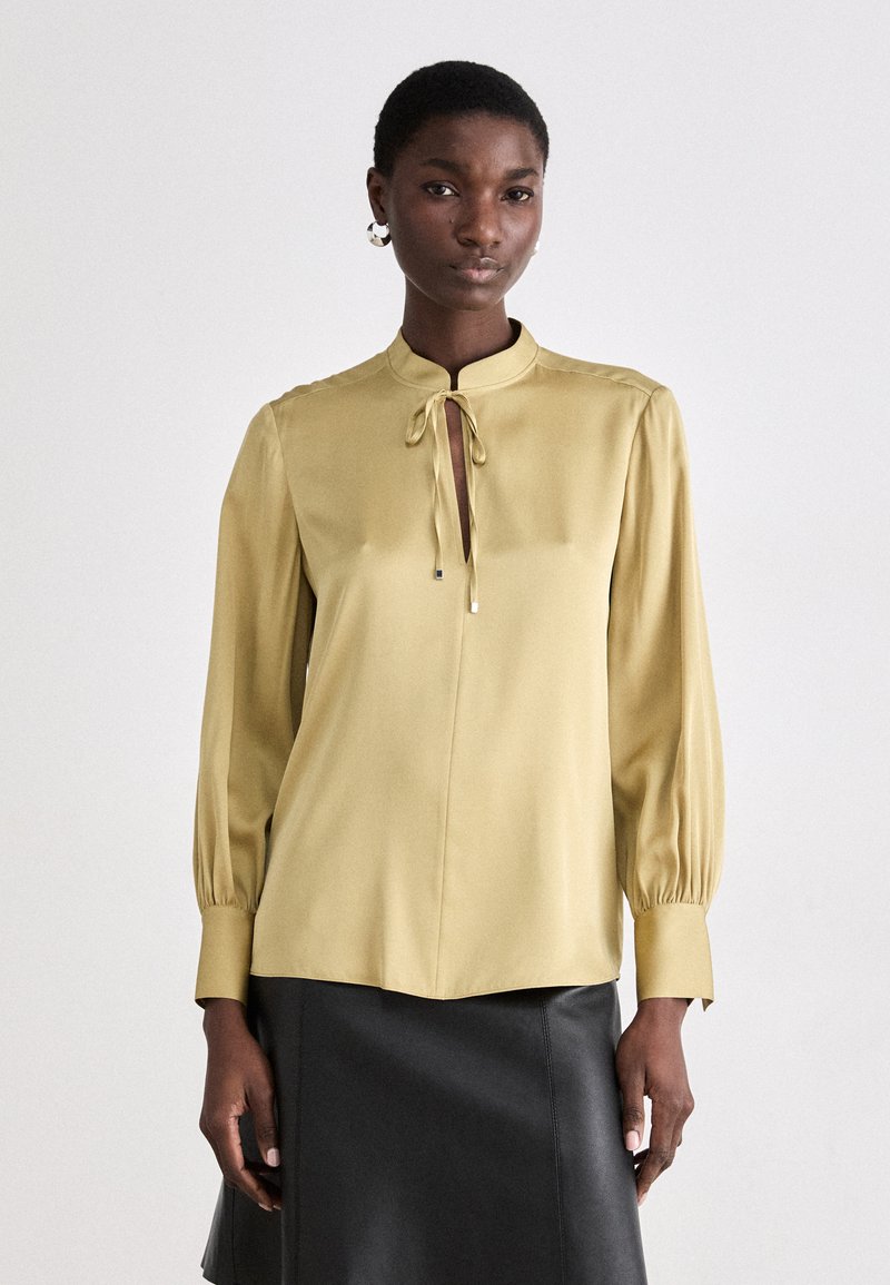 Blusa color oliva chiaro con colletto alla coreana, chiusura frontale con laccetto e maniche lunghe plissettate. Tessuto liscio con una vestibilità comoda, abbinata a una gonna di pelle nera.