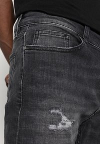 Černé džínové kalhoty s vybledlým vzhledem, s popraskanými detaily a klasickým designem pěti kapes. Kartáčovaný povrch s mírným leskem.