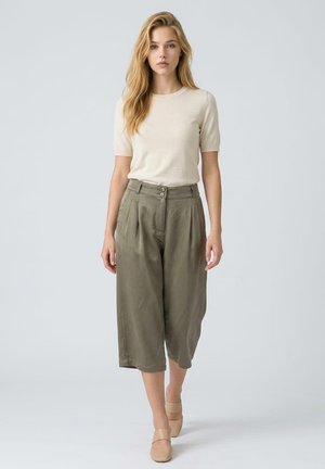 Joven mujer con cabello rubio largo que lleva una camiseta beige de manga corta, pantalones cortos anchos de color verde oliva y zapatos beige sin cordones caminando hacia adelante.