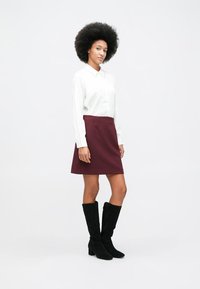 Woman with afro hair wearing a white long-sleeve blouse, maroon skirt, and black knee-high boots, standing against a plain white background.