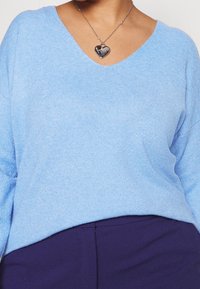 Lichtblauwe, zachte gebreide trui met een V-hals en lange mouwen, gecombineerd met een paarse onderkant en een hartvormige zilveren ketting.