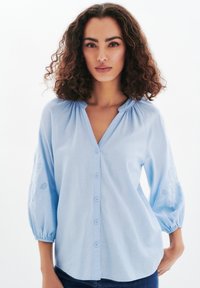 Blusa azzurra chiara con scollatura a V, chiusura con bottoni e maniche a soffietto, caratterizzata da ricami floreali e motivi aperti sul tessuto.