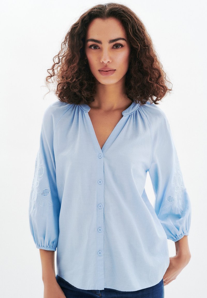 Blusa azzurra chiara con scollatura a V, chiusura con bottoni e maniche a soffietto, caratterizzata da ricami floreali e motivi aperti sul tessuto.