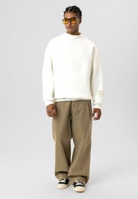 Witte oversized sweatshirt, beige wijde pantalon en zwarte sneakers. Model draagt gele zonnebril en een subtiel logo op de mouw.