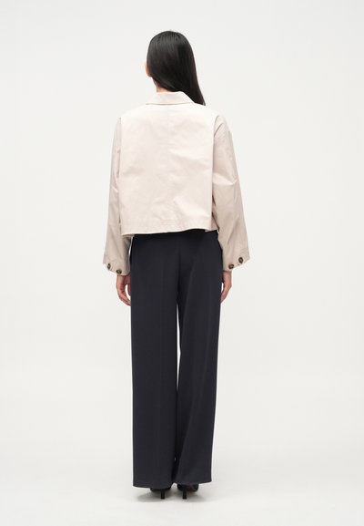 Ελαφρώς μπεζ κοντό σακάκι με γιακά, φαρδιά μανίκια και κουμπιά ως διακοσμητικά, συνδυασμένο με σκούρο μπλε παντελόνι με φαρδιά μπατζάκια.