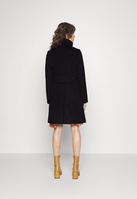 Zwarte wollen jas met een getailleerde taille en hoge kraag. Knopen aan de achterkant, gecombineerd met een oranje jurk en beige, chunky hakken laarzen.