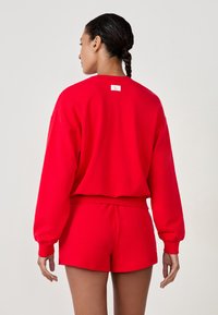 Completo di felpa rossa e pantaloni corti realizzato in tessuto morbido. La felpa ha maniche lunghe, polsini e vita a costine, con un'etichetta sul collo.