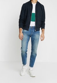 Marinblå bomberjacka med dragkedja fram, vit t-shirt med en grön grafik, ljusblå slim-fit jeans och vita sneakers.