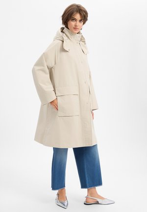 Vrouw die een beige regenjas met capuchon draagt, blauwe cropped jeans en zilveren puntige slingback platte schoenen, staand met hand in zak.