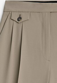 Pantalon beige avec un pli et un détail de patte en tissu boutonnée à la taille, montrant une partie de l'avant et de la zone de poche.