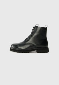 Bota de tobillo de cuero negro con punta redondeada, parte delantera con cordones y suela de goma texturizada. Presenta una lengüeta en el talón y costuras en contraste.
