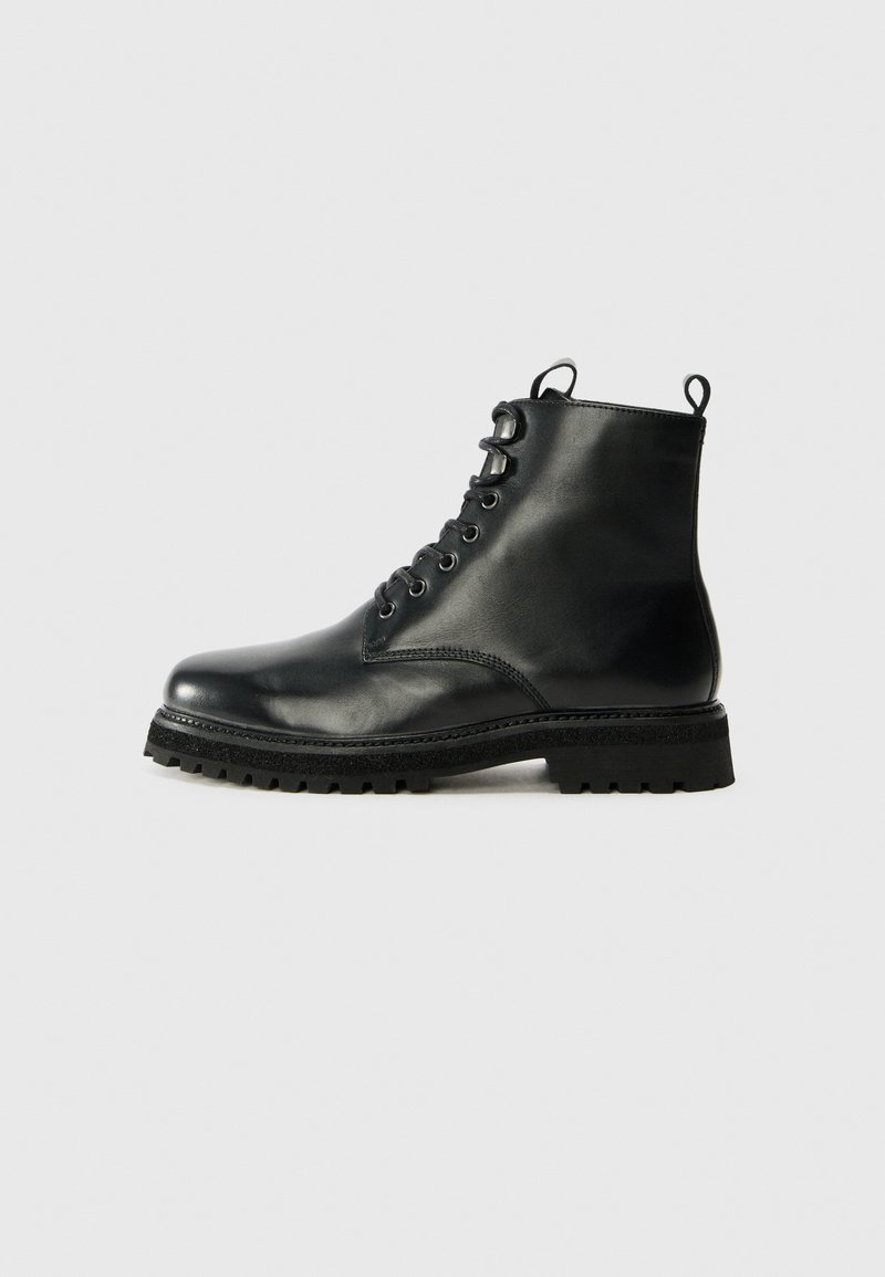 Bota de tobillo de cuero negro con punta redondeada, parte delantera con cordones y suela de goma texturizada. Presenta una lengüeta en el talón y costuras en contraste.