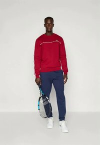 Röd tröja med vit accent, parat med marinblåa sweatpants. Modellen håller en tennisracket. Sneakers synliga. Enkel, sportig design.