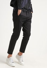 Czarne, dopasowane spodnie o gładkiej fakturze, z bocznymi kieszeniami i zwężającymi się nogawkami, w zestawieniu z białymi trampkami.