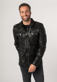 Giacca di pelle nera con chiusura a zip, due tasche sul petto, superficie texturizzata, spallini e chiusure a pressione. Indossata sopra una camicia nera.